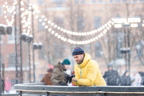 Portrait of joyful bearded man in yellow jacket, black hat standing near fence on ice rink and using smart phone, outdoors at snowy day/Winter time concept/ Weekend activities outdoor in cold weather