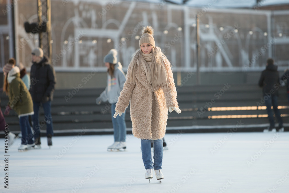 Full length portrait of young woman with blonde hair in fur coat, beige hat, scarf and blue jeans on ice rink, outdoors at sunny winter weather/ Weekends activities outdoor in cold weather/
