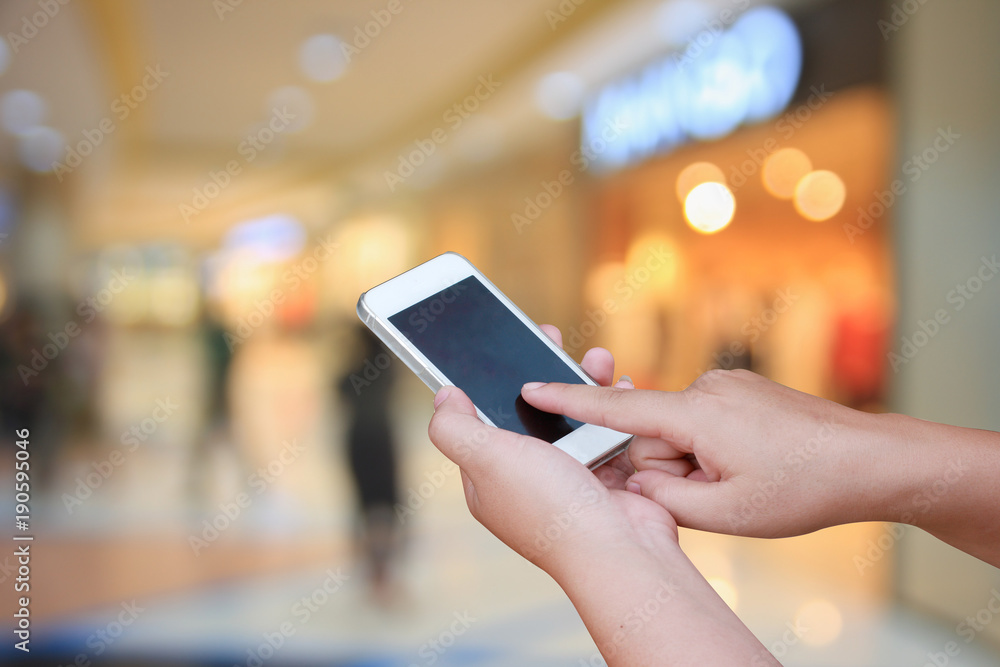 hand hold and touch screen smart phone, on blurred photo of department store shopping mall center and people background.