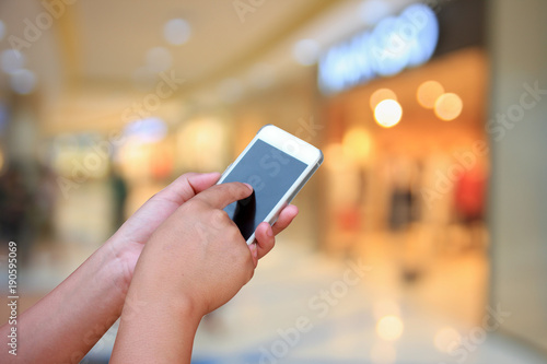 hand hold and touch screen smart phone, on blurred photo of department store shopping mall center and people background.