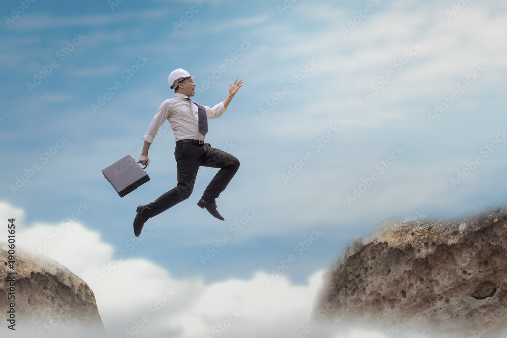 Businessman with helmet jumping over gap on the rock with sky background, obstacle and problem gap concept Businessman with helmet jumping over gap on the rock with sky background, obstacle and problem gap concept