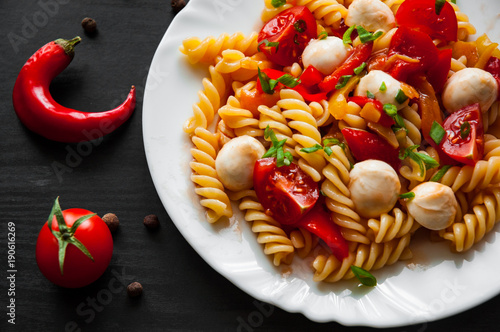 fusilli pasta salad with tomato, pepper and mozzarella balls in plate on dark wooden background