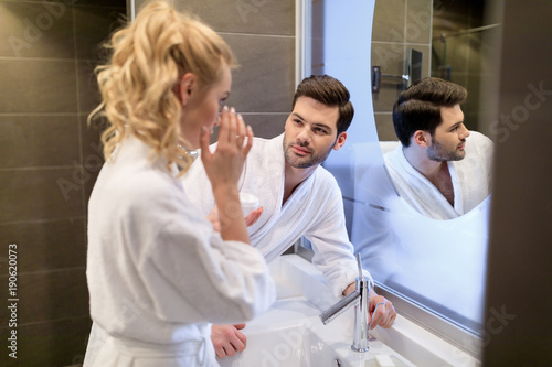 boyfriend looking how girlfriend applying cream on face in bathroom