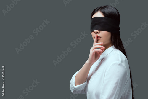 young woman with black blindfold gesturing for silence isolated on grey