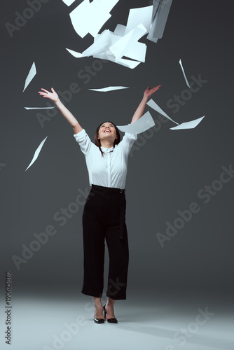 full length view of happy young businesswoman throwing papers on grey photo