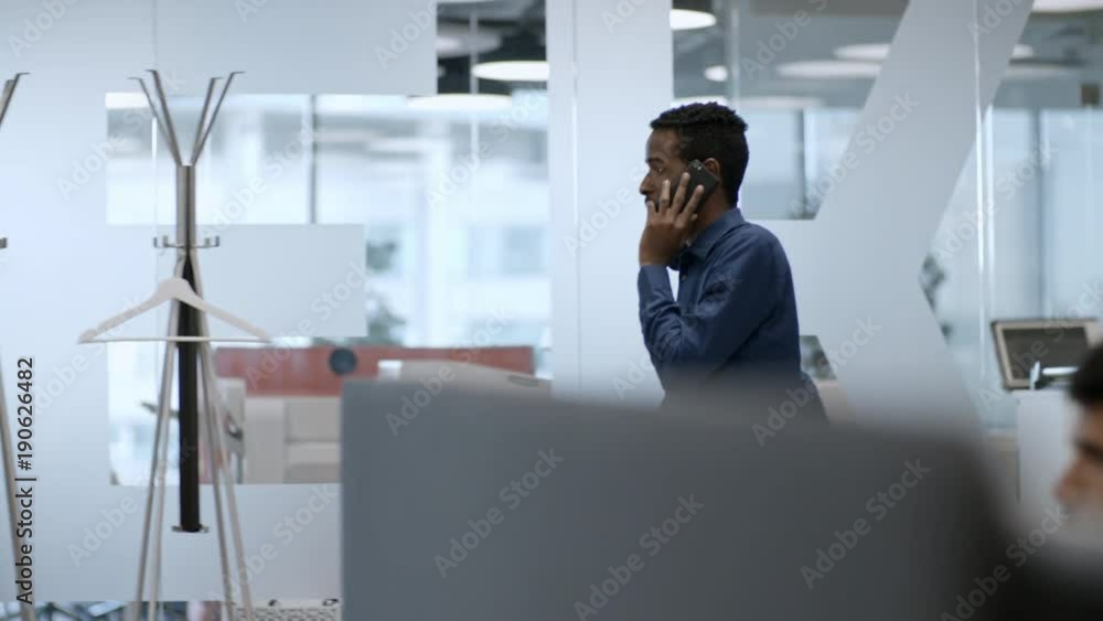 Side view of young African manager walking through modern office and speaking on mobile phone Side view of young African manager walking through modern office and speaking on mobile phone