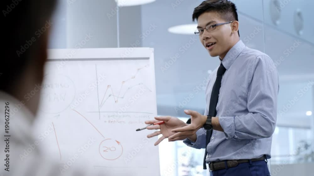 Young Asian business coach explaining scheme on whiteboard and talking with office workers while giving presentation in meeting room