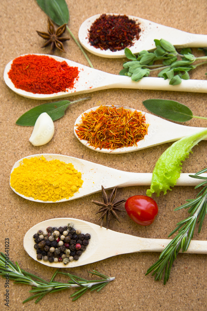 Cooking hot spicy meal. A set of four different spices in wooden spoons, fresh cherry tomatoe, garlic, rosemary and salvia, corkwood background, top view