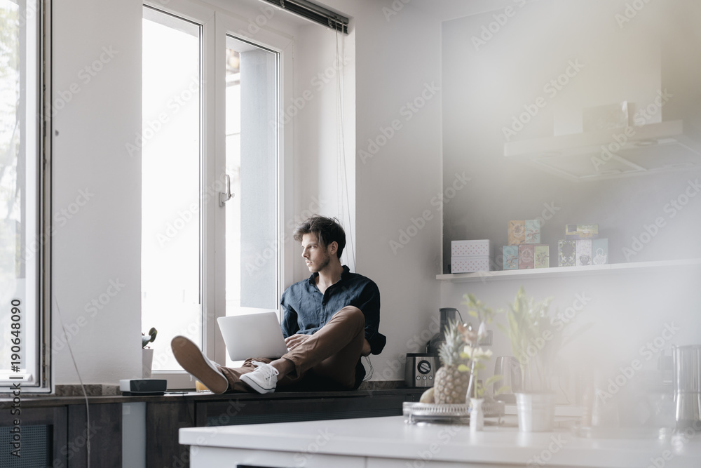 Young man with laptop sitting on window sill in a loft looking out of the window Young man with laptop sitting on window sill in a loft looking out of the window