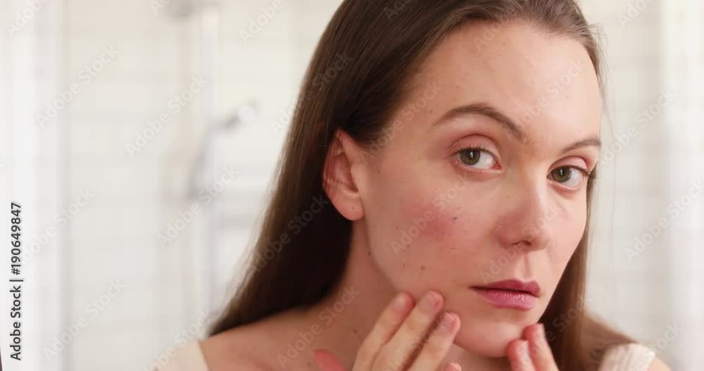 Young beautiful woman touching skin in bathroom. Unhappy girl standinglooking in the mirror, checking dry irritated skin. Morning skincare routine Young beautiful woman touching skin in bathroom. Unhappy girl standinglooking in the mirror, checking dry irritated skin. Morning skincare routine