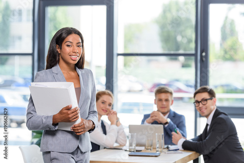 Confident businesswoman with folder in hands in front of his colleagues in modern office