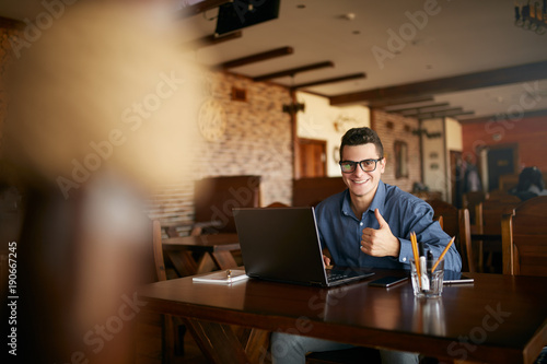 Young businessman looking at camera with laptop in cafe. Smiling man in stylish glasses and smart casual shirt doing his work on project and showing thumb up sign. Smart casual wear. Business theme.