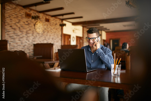 Young freelancer working with laptop and talking on cellphone with client in cafe. Handsome caucasian businessman in glasses conducts negotiations via phone call. Multitasking business concept.
