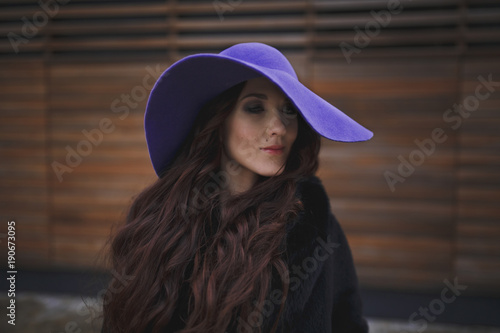 Beautiful smiling charming young woman with long hair in a ultraviolet color hat is on the background of wooden wall. Luxurious brown hair waving from the wind. The gaze is directed down