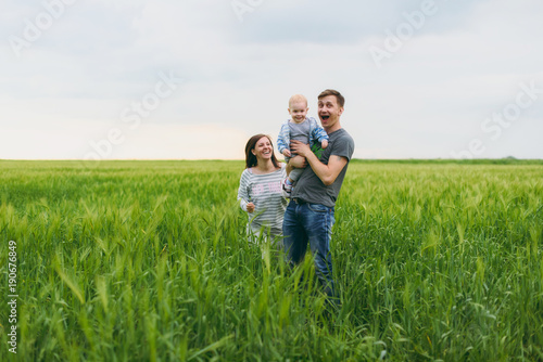 Joyful man, woman walk on green field background, rest, have fun, play, stand with little cute child baby boy. Mother, father, little kid son. Family day 15 of may, love, parents, children concept.