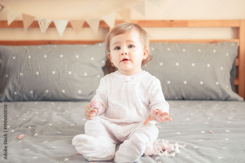 Portrait of cute adorable Caucasian blonde smiling baby girl in white onesie sitting on bed in bedroom and holding pink flower rose. Happy childhood lifestyle concept