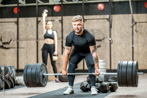Handsome athletic man in black sports wear lifting up a heavy burbell with woman training on the background in the crossfit gym