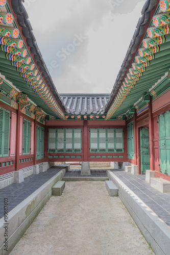 Looking down the middle of a historical building with colorful design and architectural details, in the middle a strip of dirt and the sky above, at Gyeongbokgung Palace, South Korea