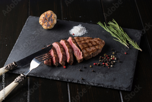 Sliced prime ribeye steak on black stone plate. Medium degree of steak doneness. With rosemary and peppers. Fork and knife.