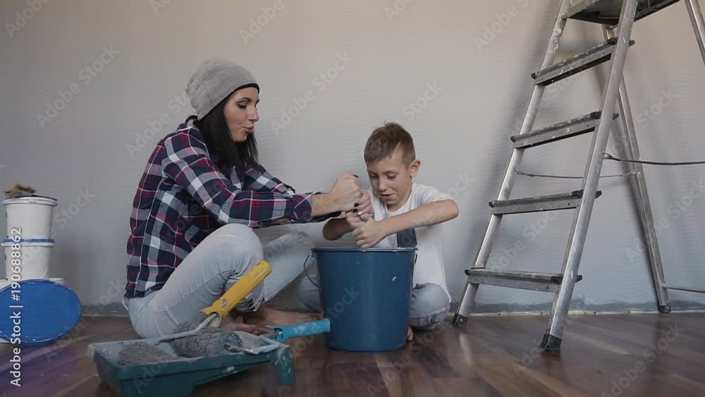 A young mother is having fun sits on the floor near the wall and holds roller in her hand, and her son sits next to her and mixes the paint that is in a bucket mixer A young mother is having fun sits on the floor near the wall and holds roller in her hand, and her son sits next to her and mixes the paint that is in a bucket mixer