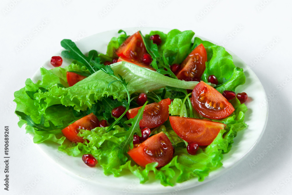 Salad on a white plate on a white background. Cherry tomatoes, arugula and pomegranate seeds on fresh lettuce leaves. Close-up.