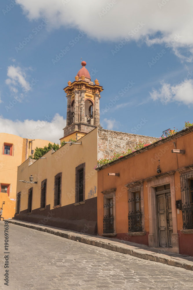 A steep cobblestone street with colorful buildings and a church tower, in San Miguel de Allende