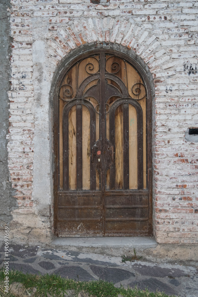 An old beautiful ornate arched door, with a white washed brick wall and brick frame work around the arched door, and a stone walkway, in San Miguel de Allende