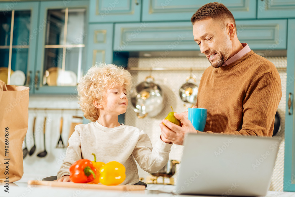 Healthy food. Positive nice handsome man standing in the kitchen and talking to his son while taking a pear from his hands Healthy food. Positive nice handsome man standing in the kitchen and talking to his son while taking a pear from his hands