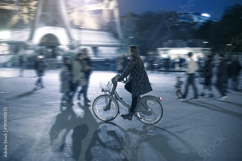 A beautiful young smiling woman riding her rental bicycle on a crowded city street during the night in front of the Eiffel Tower, Paris, France. © tannujannu