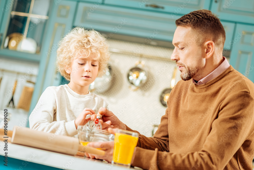Time with family. Nice positive caring father sitting together with his son and helping him to beat eggs while cooking together Time with family. Nice positive caring father sitting together with his son and helping him to beat eggs while cooking together