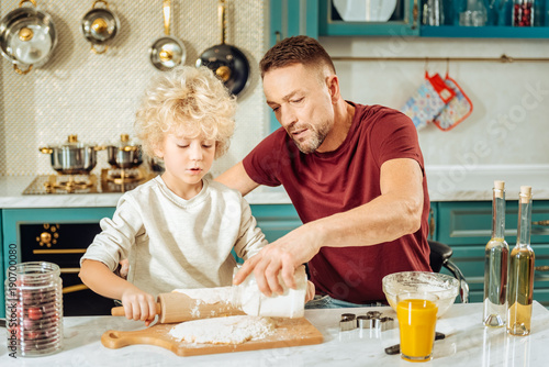In the kitchen. Handsome positive pleasant man sitting together with his son and holding a jar with flour while helping him in the kitchen