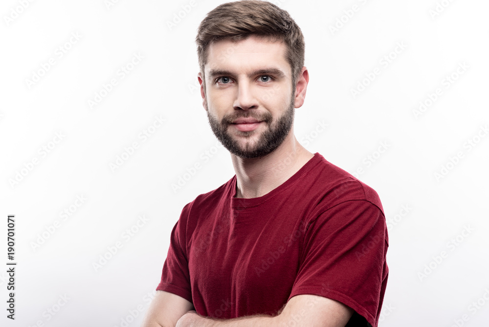 Powerful position. The portrait of a charming young man in a burgundy t-shirt standing half-turned and folding his arms across his chest while posing isolated on a white background Powerful position. The portrait of a charming young man in a burgundy t-shirt standing half-turned and folding his arms across his chest while posing isolated on a white background