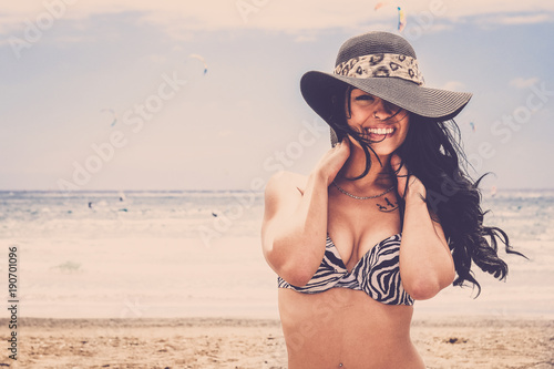 young smile beautiful woman smile at te camera on the beach in Tenerife. Long black hair touching hre shoulder back. Big grey hat and kitesurfers on the back. photo