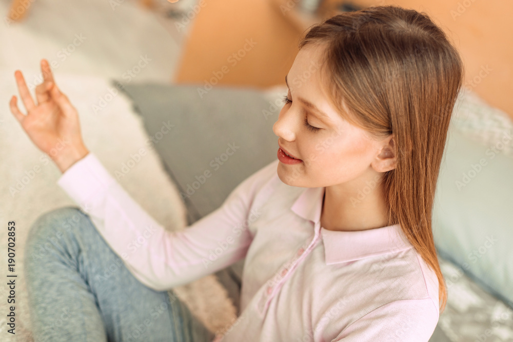 Inner peace. Beautiful teenage girl sitting cross-legged in her room and folding her fingers in mudra while meditating Inner peace. Beautiful teenage girl sitting cross-legged in her room and folding her fingers in mudra while meditating