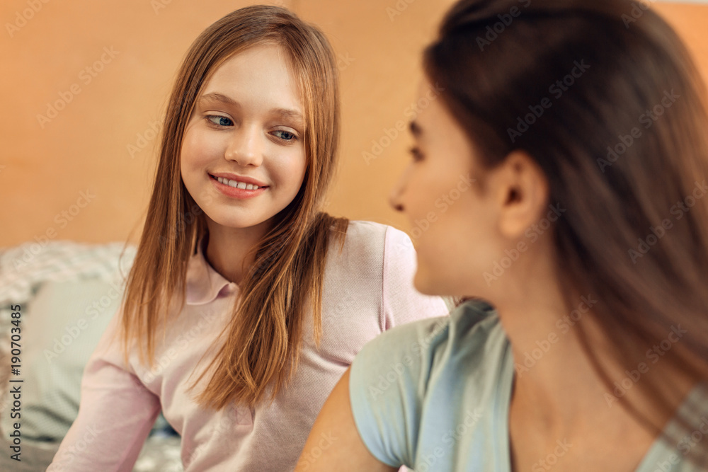 Admiring look. Cute teenage girl sitting on the bed next to her elder sister and looking at her with admiration while smiling