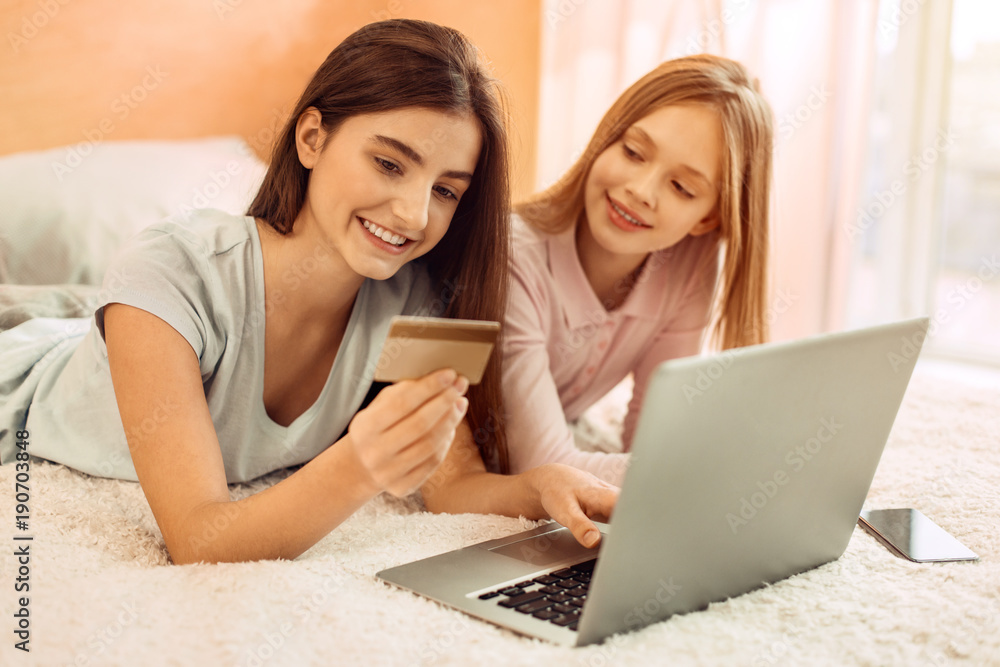 Fast and convenient. Pleasant teenage sisters lying on the bed and doing online shopping together while the older girl paying with her bank card