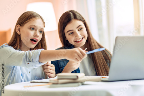 Check it out. Pleasant teenage girl sitting at the table next to her sister and pointing at a laptop screen, attracting her sisters attention © Viacheslav Yakobchuk