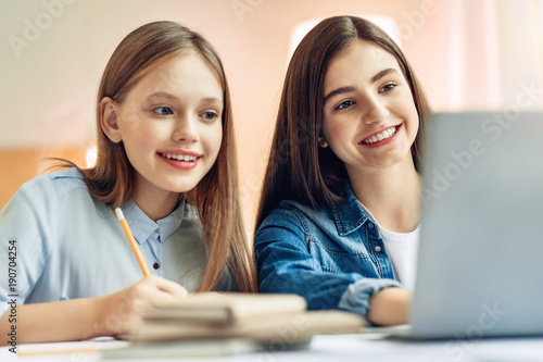 Helping each other. Beautiful teenage girls sitting at the table in the living room and studying together while reading the tasks from their laptop