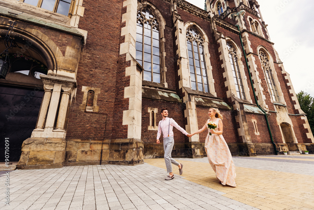 A beautiful stylish boy holds his girlfriend's hand and they run along the background of a beautiful building