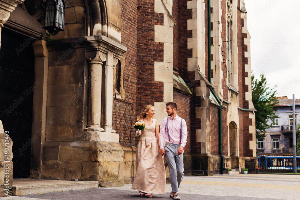 the girl in a long dress holds a bouquet and her boyfriend next to her and they walk together at the entrance to a beautiful brick building
