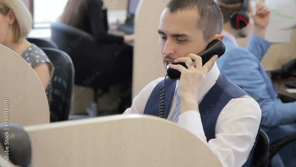The portrait of the manager who talks on phone with client in the big office among the colleagues. The professional wears dark vast, blue tie and silver fingerring and sit at the table with separation The portrait of the manager who talks on phone with client in the big office among the colleagues. The professional wears dark vast, blue tie and silver fingerring and sit at the table with separation