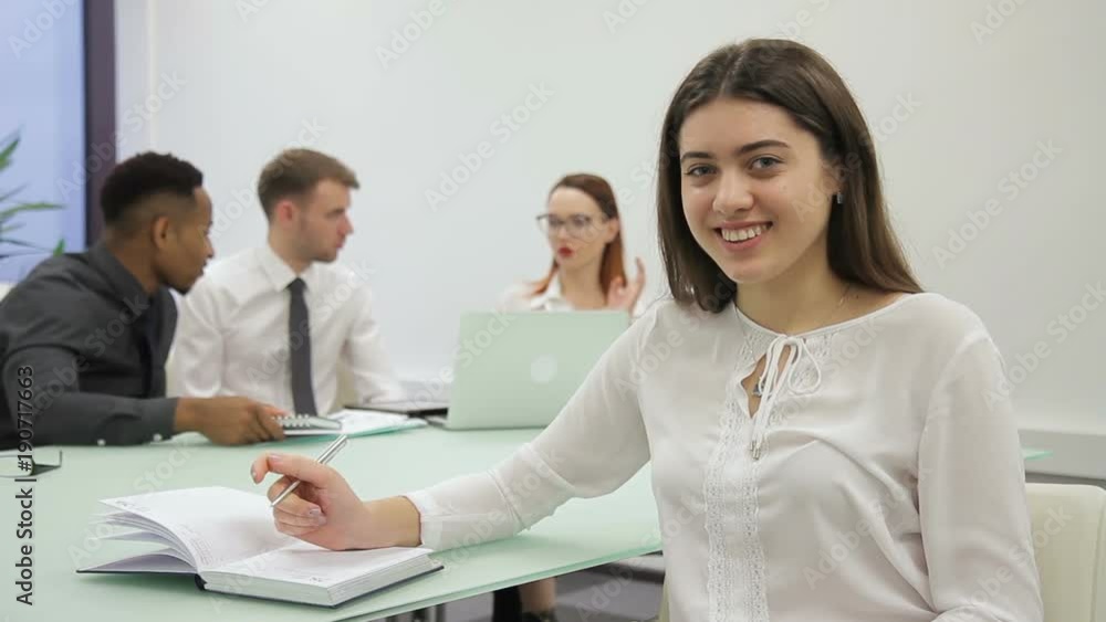 Young woman writes then looking ahead, sitting in modern office. Attractive female is at table, takes notes on pages of notebook with pen then distracts and looks with charming smile. Expert dressed Young woman writes then looking ahead, sitting in modern office. Attractive female is at table, takes notes on pages of notebook with pen then distracts and looks with charming smile. Expert dressed