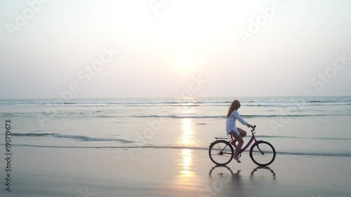 A young pretty girl riding on  her bicycle at sunset. She rushes along the seashore beautifully reflected in the water on the sand. Flying camera moves near. Aerial view.