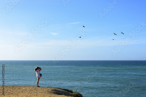 Girl on the distance  taking photos of pelicans flying over sea