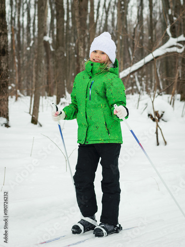 The girl in the green jacket and white hat posing on skis in the winter woods.
