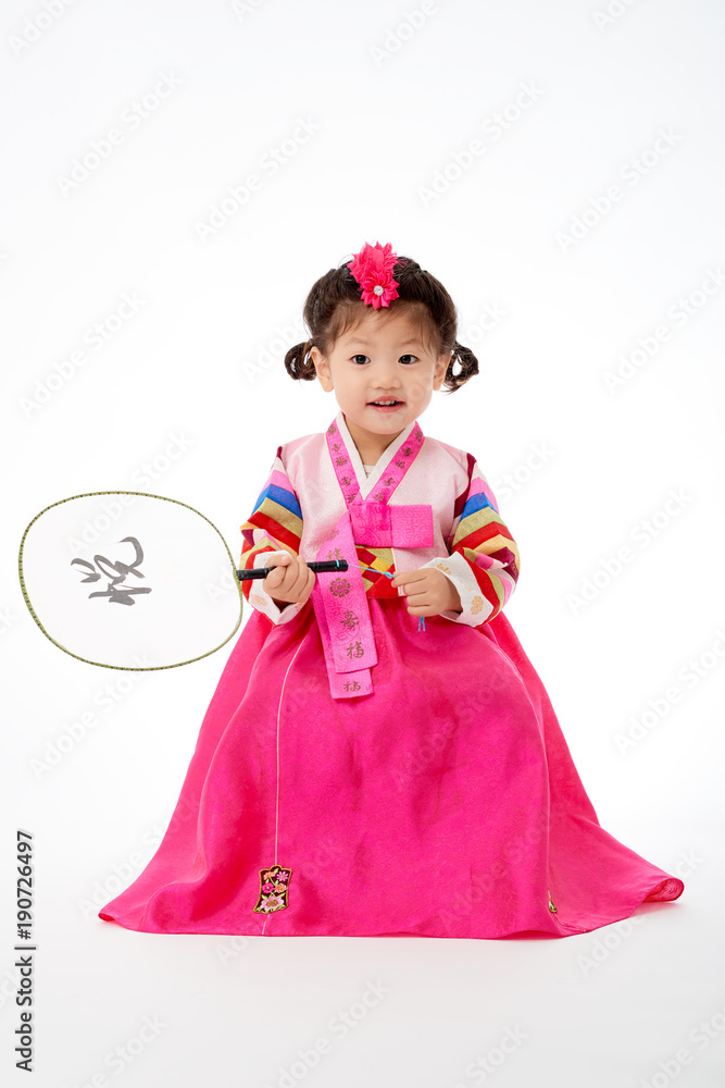 Smile Korean little girl wearing a Traditional Hanbok dress and holding a fan in white background. The characters on the dress means happiness and healthy. The character on the fan means happy.