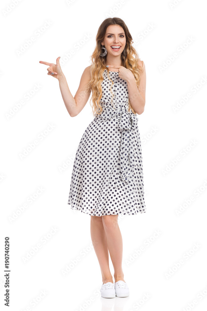 Smiling Woman In Dotted Dress Is Pointing And Looking At Camera.