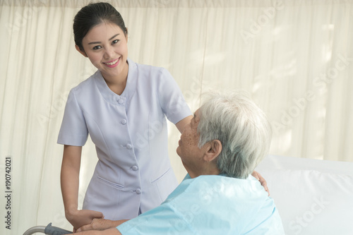 Cheerful asian nurse visiting elderly patient to check up after surgery in hospital for giving physical therapy and encouragement.