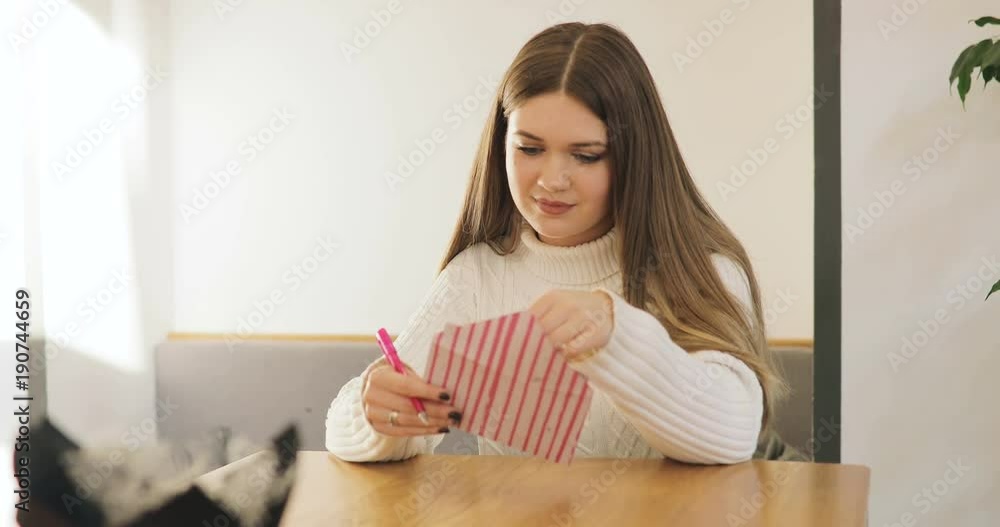 Brown-haired young women in white sweater write, smiling, get, read a Valentine's Day card and sign in reply in stylish cafe, home. Brown-haired young women in white sweater write, smiling, get, read a Valentine's Day card and sign in reply in stylish cafe, home.