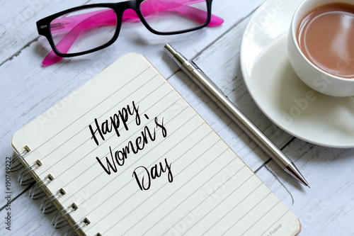 Top view of notebook written ' HAPPY WOMEN'S DAY' with a cup of coffee, pen and sunglasses on white wooden background.